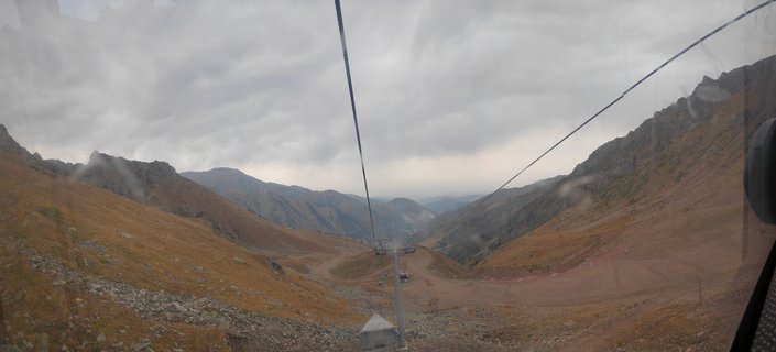Panoramic view from inside a cable car showing mountain valley with gondola lines, rocky slopes with brown vegetation, and cloudy sky at Shymbulak Resort in Kazakhstan