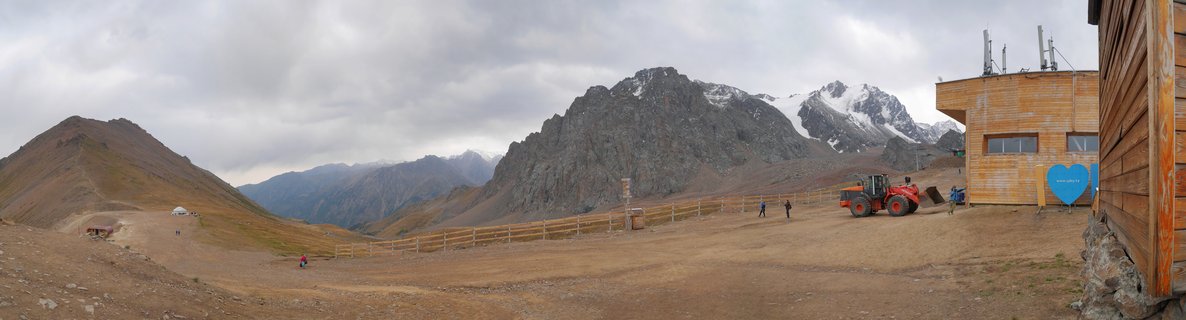 Wide mountain landscape showing rocky peaks with snow, wooden buildings, a red tractor, wooden fencing, and small figures of people walking on dirt ground under cloudy sky