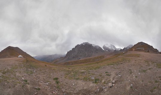 Panoramic mountain landscape showing rocky peaks, alpine meadow with sparse vegetation, a white yurt on the left hillside, and overcast cloudy sky in Kazakhstan