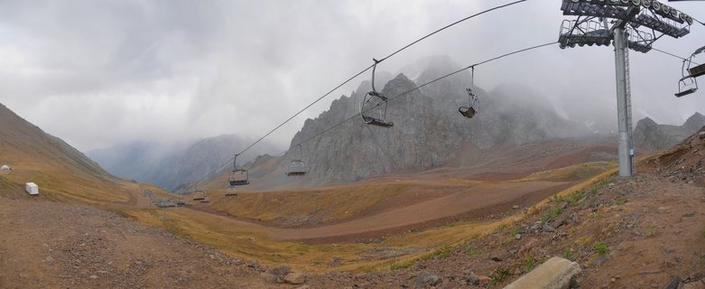 Empty chairlift chairs suspended on cables ascending a mountainside with rocky peaks shrouded in fog, brown alpine grassland below, Kazakhstan