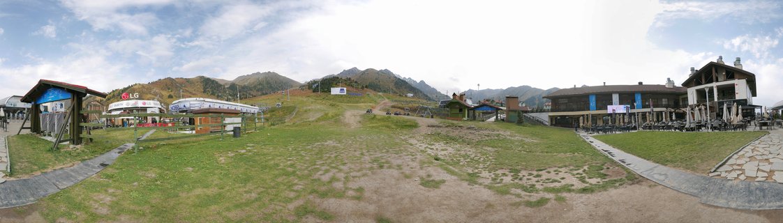 Wide panoramic view of Shymbulak Ski Resort showing gondola stations, resort buildings with outdoor seating, grassy slopes, and mountain peaks under partly cloudy sky in Almaty, Kazakhstan