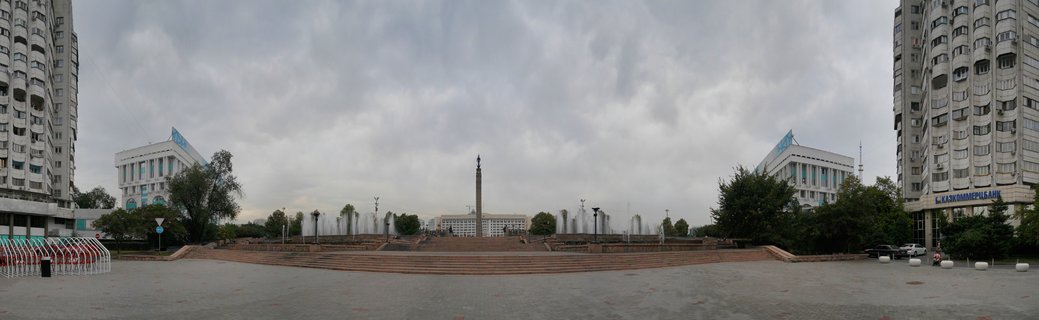 Wide panoramic view of Republic Square in Almaty showing a central obelisk monument, symmetrical fountains, tall apartment buildings on both sides, and white government buildings with blue accents under cloudy sky