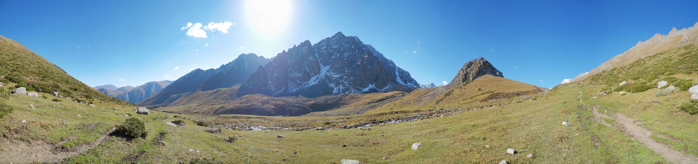 Wide mountain landscape showing rocky peaks with snow patches rising above a green alpine valley with hiking trails and scattered boulders under bright blue sky