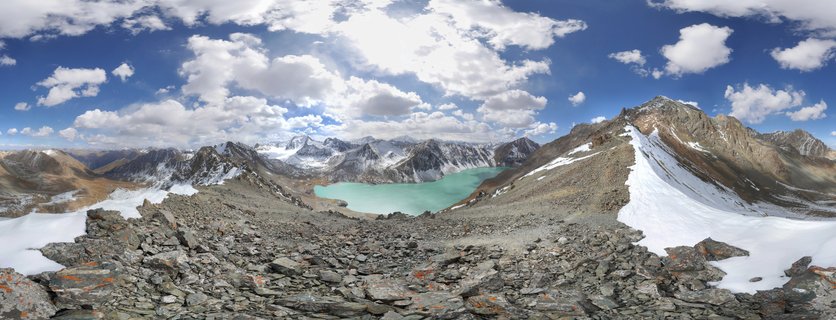 Panoramic landscape showing a turquoise alpine lake surrounded by rocky mountains with snow patches under a blue sky with white clouds in Kyrgyzstan