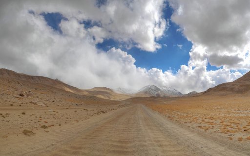 A wide dirt road leading through a barren mountain desert landscape toward distant snow-covered peaks under a cloudy blue sky in Tajikistan