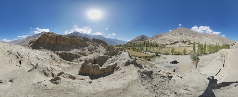 Wide panoramic landscape showing eroded beige rock formations in foreground, green valley with trees in middle distance, and brown mountain ranges under bright blue sky with visible sun