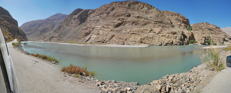 Panoramic landscape showing a turquoise river flowing through a rocky mountain valley with a dirt road along the riverbank under clear blue sky in Tajikistan