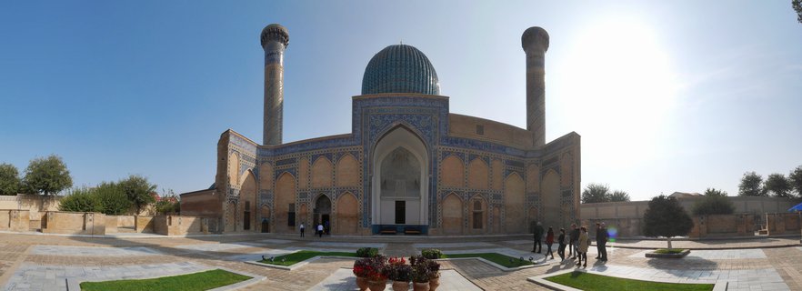 Historic Bibi-Khanym Mosque in Samarkand featuring a large turquoise dome centered between two tall minarets, with blue and gold Islamic tilework on sandstone walls, visitors walking in the paved courtyard under bright sunlight