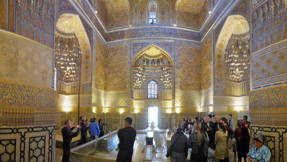 Interior view of an ornate Islamic mausoleum with blue and gold tiled walls, decorative arches, hanging chandeliers, and visitors walking among marble tombs