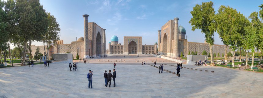 Wide panoramic view of Registan Square showing three ornate madrasa buildings with blue domes, minarets, and decorative tilework, with visitors walking across the stone plaza