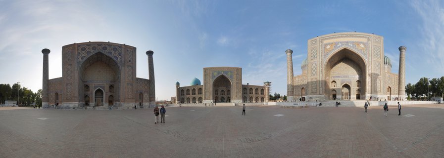 Wide view of Registan Square showing three ornate Islamic madrasah buildings with blue domes, tall minarets, and decorative tilework under a clear blue sky with tourists walking across the brick plaza