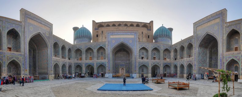 Wide courtyard view of Registan Square showing ornate blue-tiled Islamic architecture with twin turquoise domes, arched walkways, and visitors walking on stone pavement in Samarkand, Uzbekistan