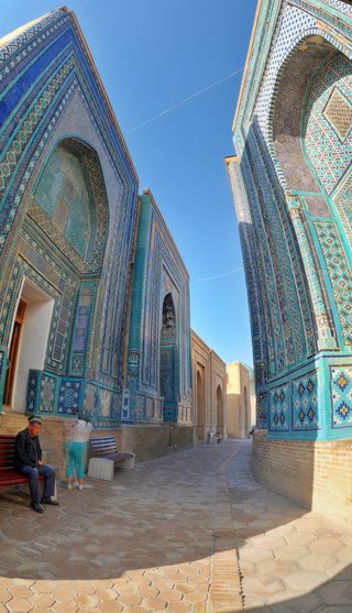 Two ornate blue-tiled mausoleum facades with Islamic geometric patterns frame a stone pathway at Shah-i-Zinda necropolis in Samarkand, with a man sitting on a bench in the foreground.