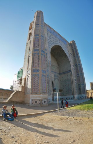 Large Islamic madrasah building with blue mosaic tilework covering the facade, featuring a massive arched entrance portal, with tourists sitting and walking in the foreground under clear blue sky