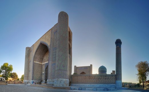 Historic Islamic madrasah with large arched entrance, ornate blue and turquoise tile mosaics, tall cylindrical minaret, and dome visible against clear blue sky at Registan Square, Samarkand