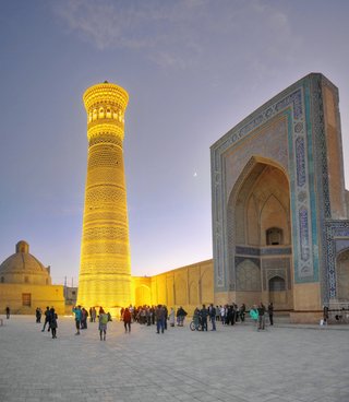 Illuminated Kalyan Minaret tower next to ornate blue-tiled mosque entrance with visitors walking in courtyard at dusk