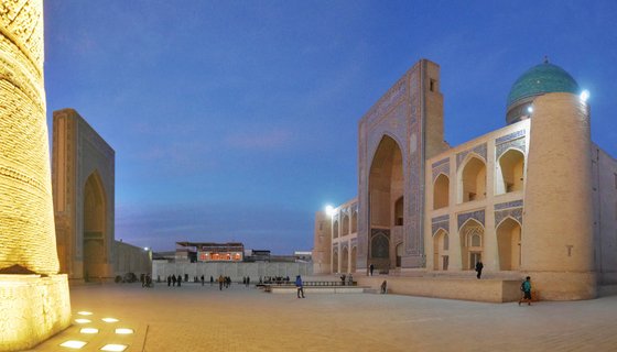 Historic Islamic architecture complex at twilight showing Kalyan Minaret and madrasah with turquoise dome, illuminated courtyard with visitors in Bukhara, Uzbekistan