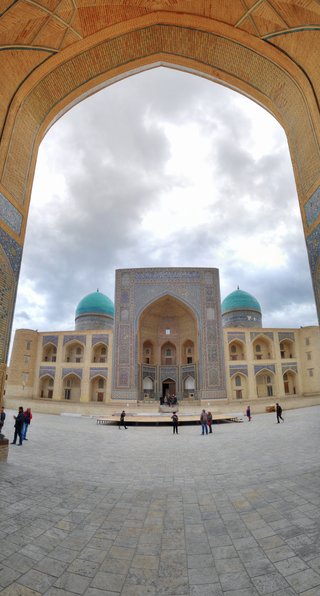 View through a large brick archway showing a historic Islamic building with two turquoise domes and ornate blue tilework in Bukhara, Uzbekistan, with visitors walking in the courtyard below
