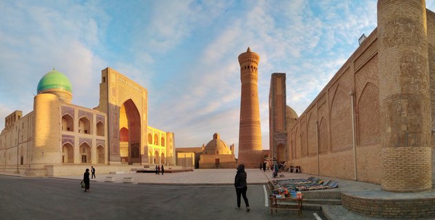 Wide view of Bukhara's Po-i-Kalyan complex showing the Kalyan Minaret, turquoise-domed Mir-i-Arab Madrasah, and historic brick buildings with people walking in the plaza during golden hour