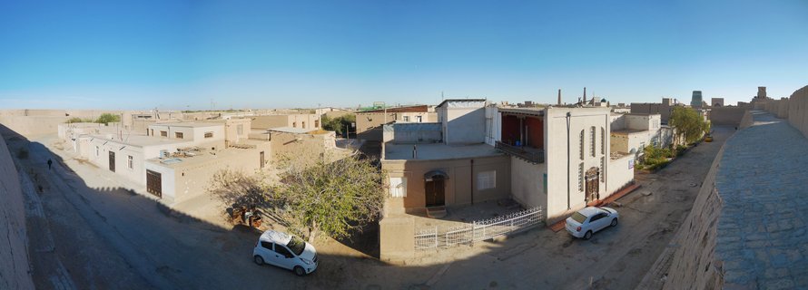 Elevated view showing traditional mud-brick buildings of Khiva's old town with flat roofs, narrow streets, parked white cars, and a minaret visible in the distance under clear blue sky