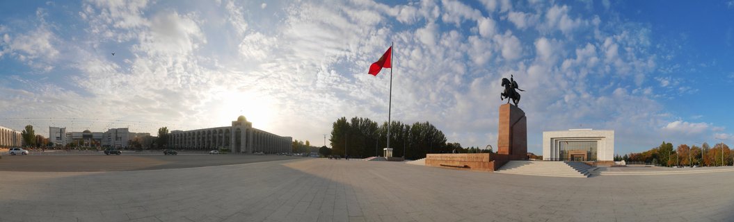Wide panoramic view of Ala-Too Square in Bishkek showing a large red flag on a tall pole, an equestrian statue on a pedestal, government buildings, and blue sky with scattered white clouds