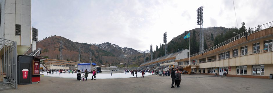 Wide panoramic view of an outdoor ice skating rink with numerous skaters on the ice, stadium bleachers on the right, tall light towers, and forested mountains with snow patches in the background under an overcast sky. A Kazakhstan flag flies on a flagpole.