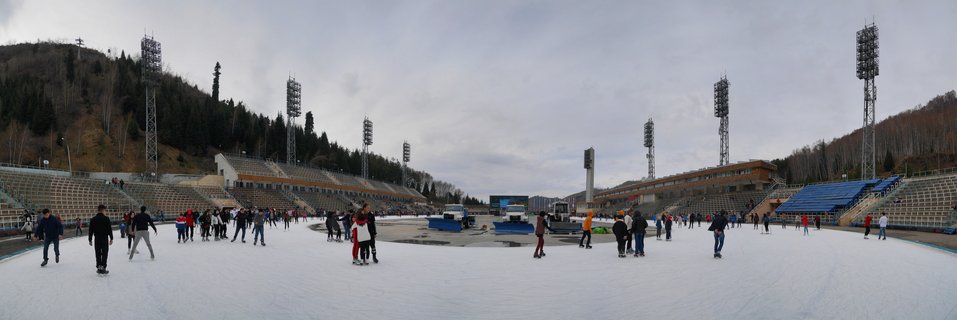 Wide view of outdoor ice skating rink with numerous skaters, stadium bleachers on both sides, tall metal floodlight towers, and tree-covered hills under cloudy sky