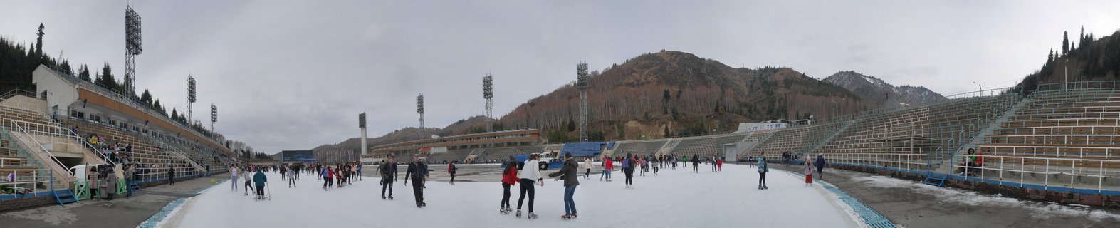 Panoramic view of outdoor ice skating rink with numerous skaters on white ice surface, flanked by concrete stadium bleachers on both sides, tall metal floodlight towers, and brown mountain slopes with evergreen trees in background under overcast gray sky