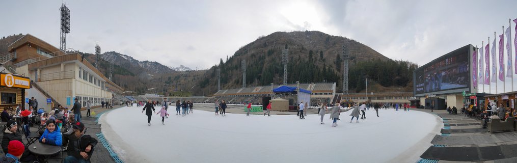 Wide panoramic view of outdoor ice skating rink with numerous people skating on white ice surface, stadium seating and buildings on sides, large mountain with pine trees in background under overcast sky