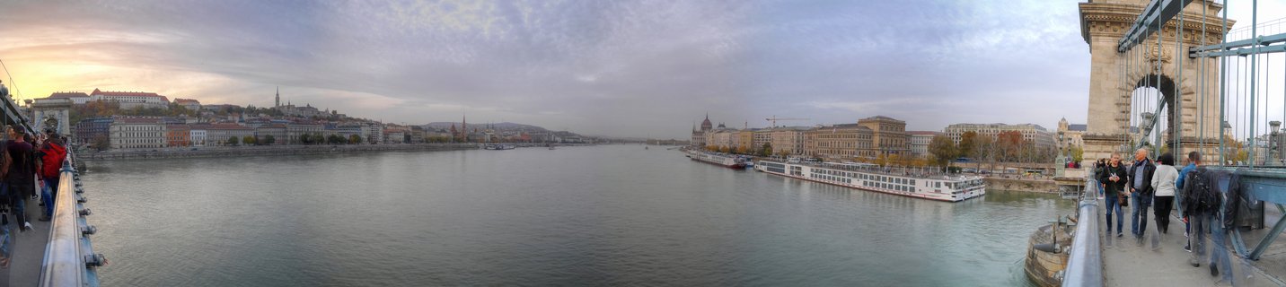Wide panoramic photograph showing Budapest's Danube River from Chain Bridge with Buda Castle on hillside left, Parliament dome in distance right, river cruise ships docked along embankment, pedestrians walking on bridge