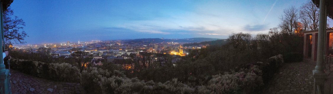 Panoramic view of Brno city at dusk with illuminated buildings, stone terrace foreground with columns and vegetation, deep blue twilight sky