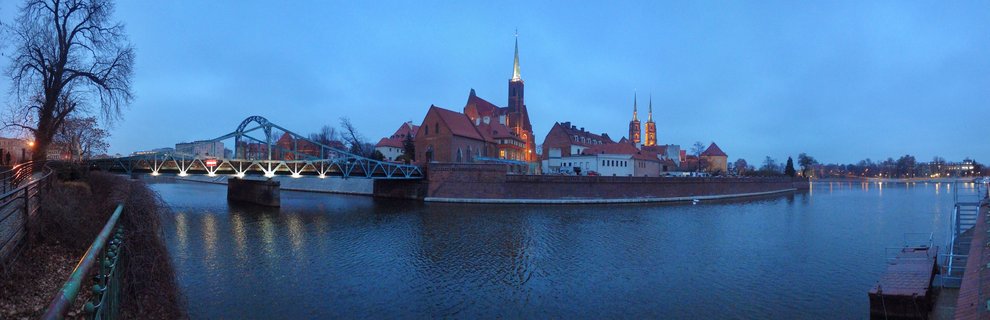 Panoramic cityscape of Wroclaw's Cathedral Island at twilight showing illuminated Gothic church spires, a blue steel bridge over the Oder River, and historic brick buildings with reflections in the water