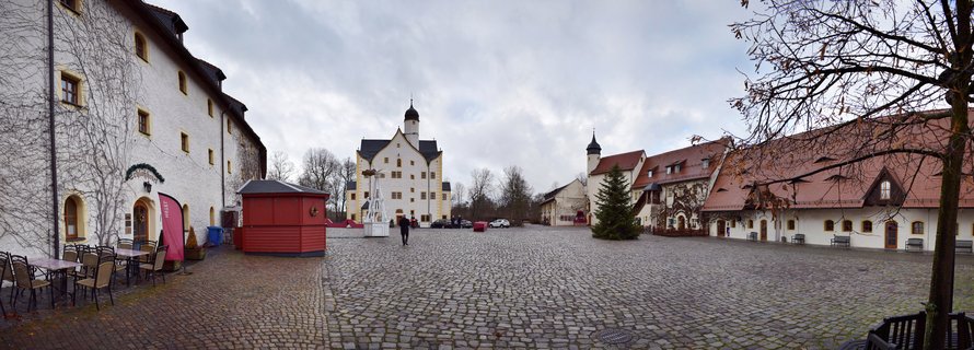 Wide panoramic view of a historic German castle courtyard with white buildings, red tile roofs, cobblestone ground, Christmas market booth and tree, overcast winter day
