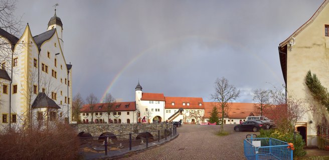 Panoramic view of a historic German castle courtyard with white buildings, red tile roofs, stone bridge, and a complete rainbow arching across a gray stormy sky