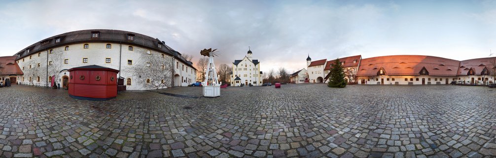 Wide panoramic view of a historic German castle courtyard with white buildings, red tile roofs, cobblestone ground, and Christmas market decorations under cloudy winter sky