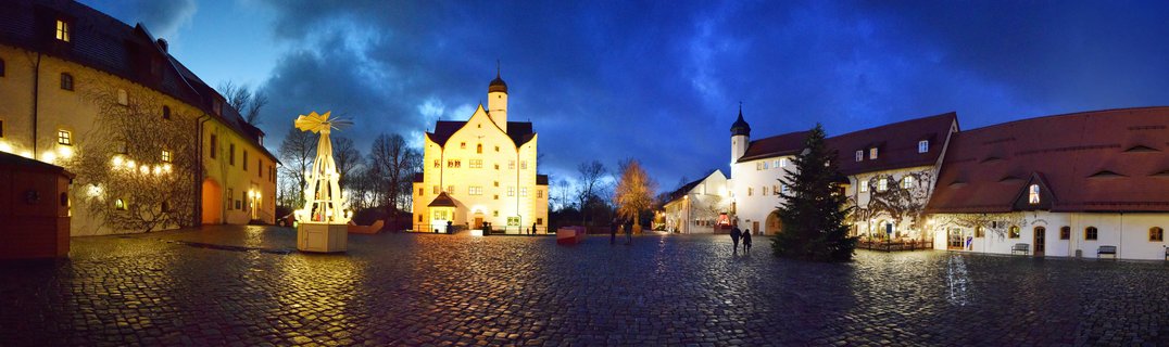 Illuminated historic castle courtyard at blue hour with Christmas pyramid, Christmas tree, and people walking on cobblestone plaza in Chemnitz, Germany