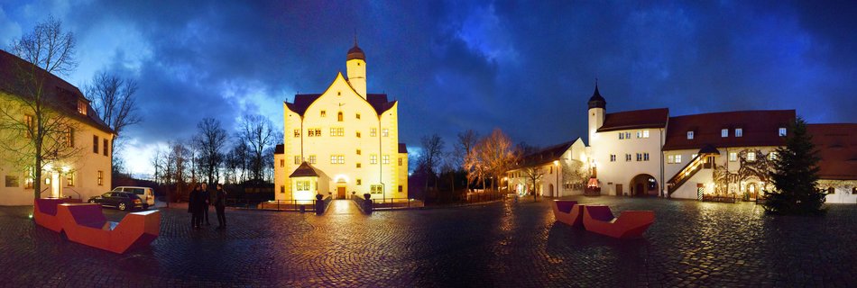 Panoramic photograph of an illuminated historic castle complex at blue hour with cobblestone courtyard, modern red benches, and visitors in Chemnitz, Germany