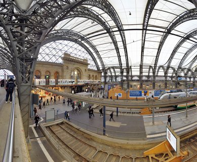 Interior view of Dresden train station showing steel arch roof structure, platforms with travelers, historic stone building facade, and modern trains at the station in Germany.