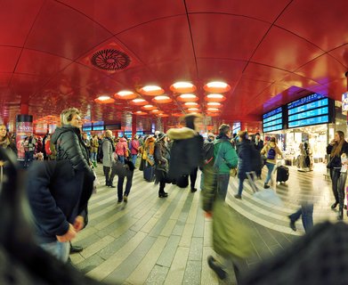 Interior view of a busy train station with a bright red ceiling featuring circular light fixtures, travelers walking with luggage, and blue digital departure boards displaying train information