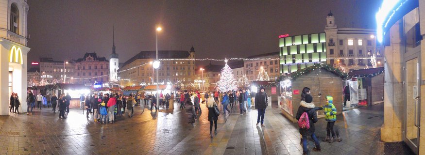 Nighttime panoramic view of a Christmas market in a European city square with illuminated Christmas tree, wooden vendor stalls, decorative string lights, crowds of people in winter clothing, historic buildings, and a clock tower visible in the background
