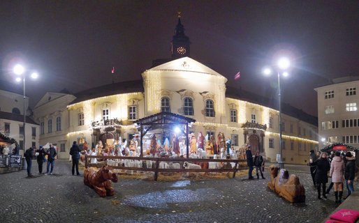 Night view of outdoor nativity scene with wooden figures in town square, baroque building with clock tower decorated with Christmas lights in background, people walking on cobblestone pavement