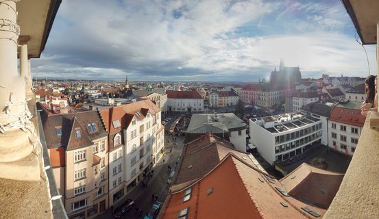 Aerial view of Prague showing red-tiled rooftops, historic buildings, and St. Vitus Cathedral spires in the distance under cloudy sky with stone architectural elements framing the edges