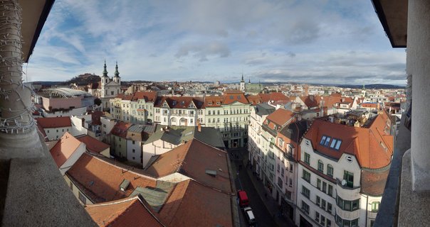 Elevated view of Prague showing red-tiled roofs, historic buildings with baroque church towers, and Prague Castle on a hill under cloudy sky