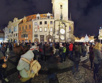 Night view of Prague's Old Town Square showing the illuminated Astronomical Clock tower, colorful historic buildings, Christmas tree, and crowds of people on cobblestone pavement