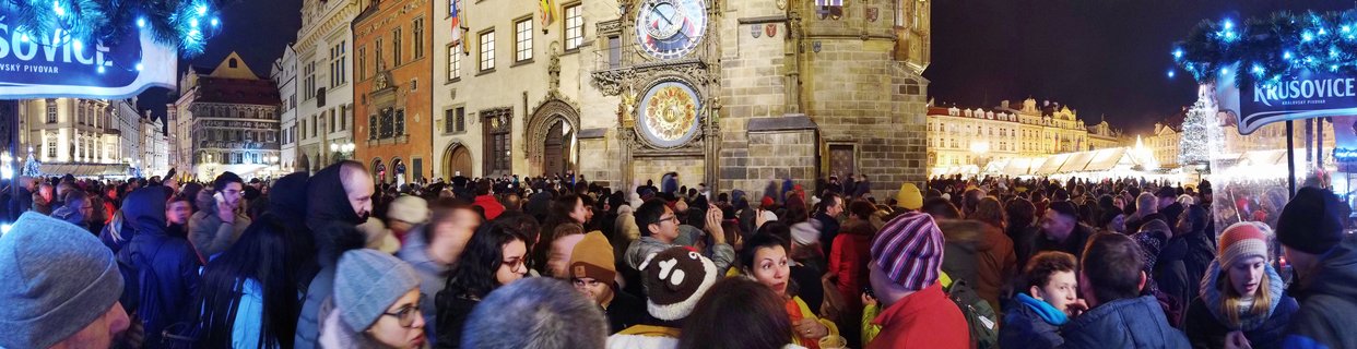 Large crowd of people in winter clothing gathered in Prague Old Town Square at night during Christmas market, with the illuminated Astronomical Clock visible on the Old Town Hall tower and festive market stalls with white tents and decorated Christmas trees with blue lights
