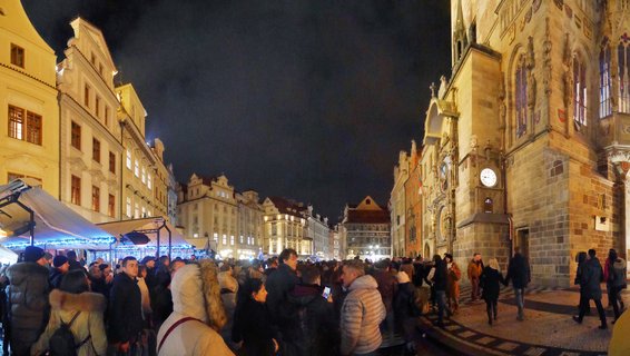 Night view of Prague's Old Town Square filled with people at Christmas market stalls, with the illuminated Astronomical Clock tower visible on the right side