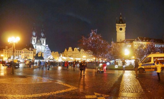 Night view of Prague's Old Town Square during Christmas market with illuminated Gothic church towers, decorated Christmas tree, market stalls with white tents, crowds of people on cobblestone pavement, and historic buildings with warm artificial lighting