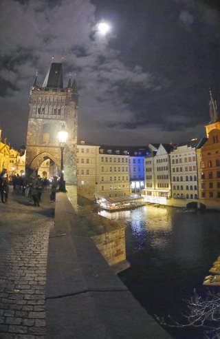 Night view of Prague's Charles Bridge showing the Old Town Bridge Tower, illuminated historic buildings along the Vltava River, pedestrians on cobblestone path, and a bright moon in cloudy sky