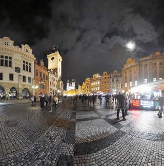 Night view of Prague Old Town Square with illuminated historic buildings, Astronomical Clock Tower, cobblestone pavement, Christmas market stalls, and people walking under a cloudy moonlit sky