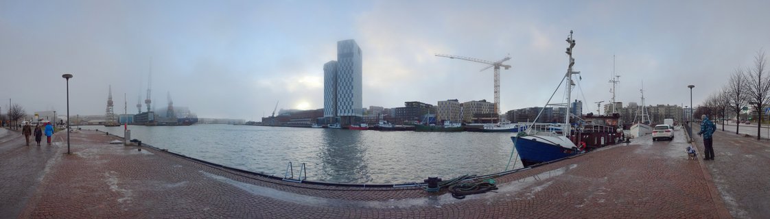 Wide panoramic photograph of Tallinn harbor showing cobblestone waterfront promenade with docked blue fishing boat, modern glass high-rise building, construction cranes, and people walking along the water on an overcast winter day